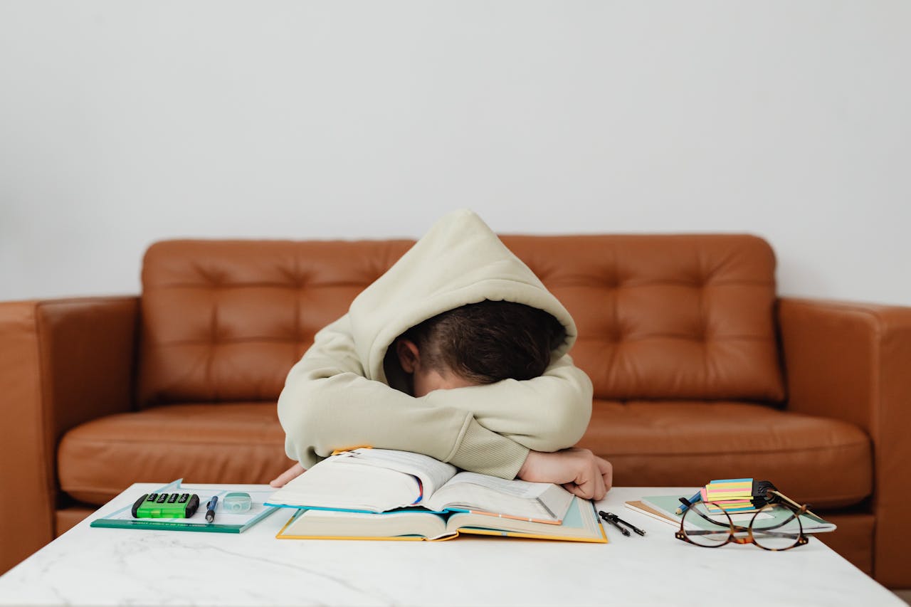 Child in hoodie leans over books, exhausted while studying on a couch.